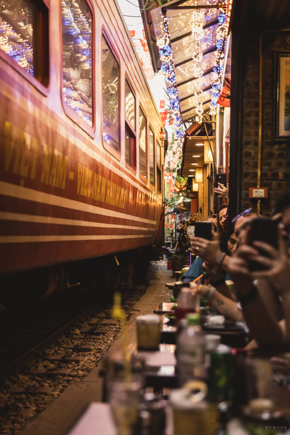 Seitliche Nahaufnahme eines Zuges, der durch die Hanoi Train Street fährt während am Rand Menschen sitzen und fotografieren