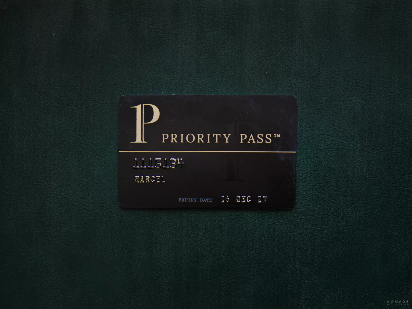 Eine schwarze Priority Pass Mitgliedskarte mit goldener Aufschrift, die flach auf einer dunkelgrünen, lederartigen Oberfläche liegt.