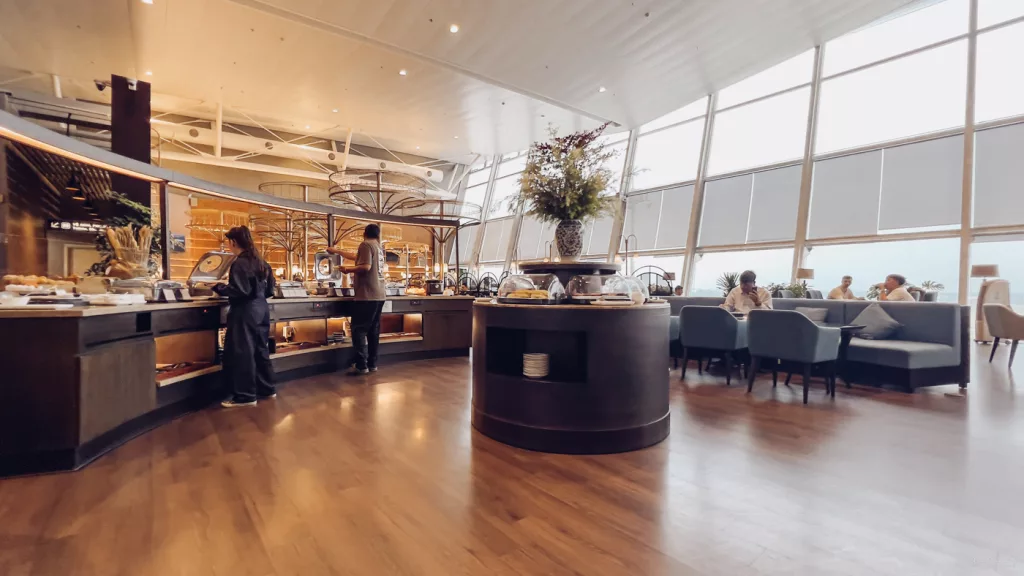 Ein weitläufiger Buffet-Bereich in einer Flughafen-Lounge mit einer großen Auswahl an Speisen und einer hellen Fensterfront, die den Blick auf das Rollfeld ermöglicht