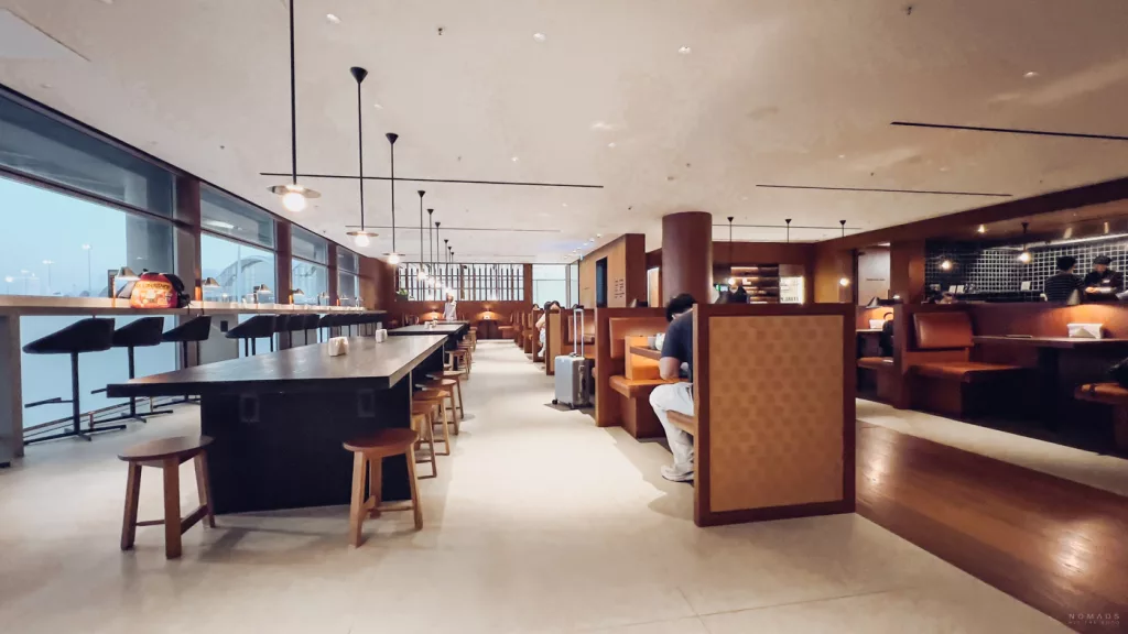 Ein moderner, minimalistisch gestalteter Essbereich in einer Lounge mit langen Holztischen, Pendelleuchten und komfortablen Sitznischen für Reisende.