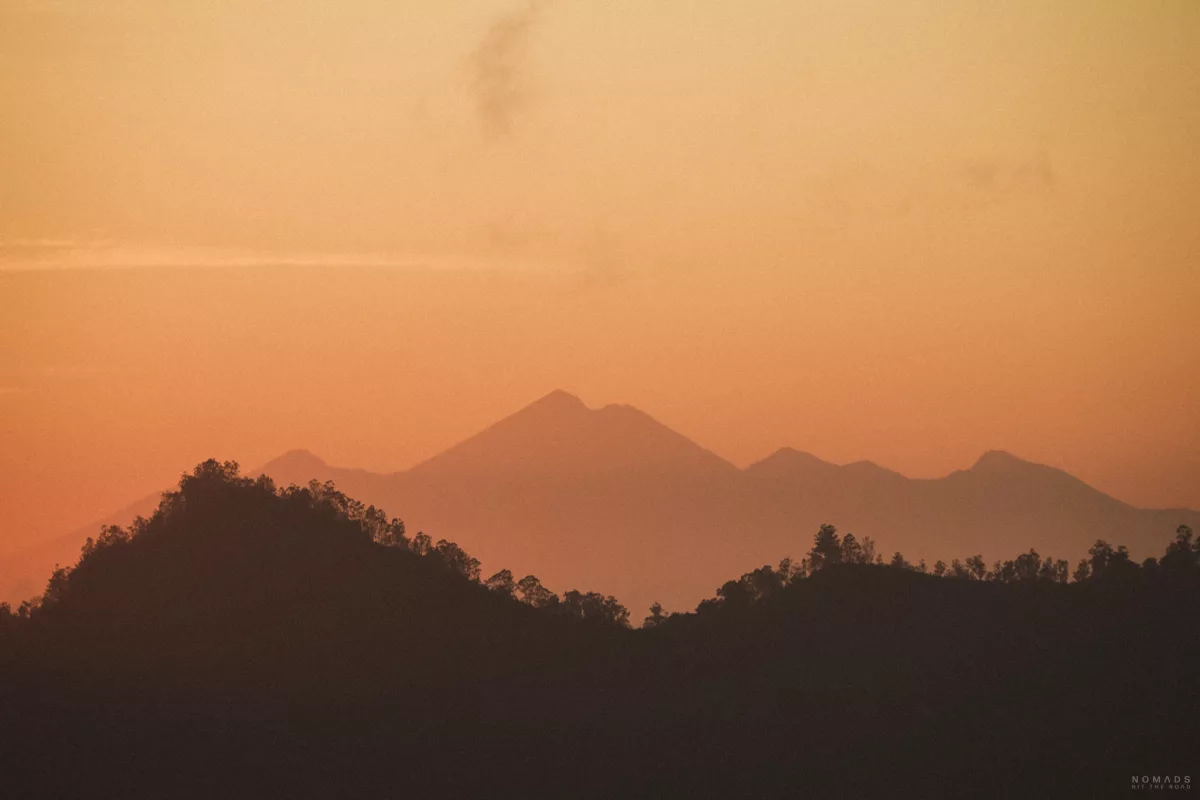 Orangfarbener Himmel bei Sonnenaufgang am Mount Batur mit Bergkette im Hintergrund und Bäumen davor