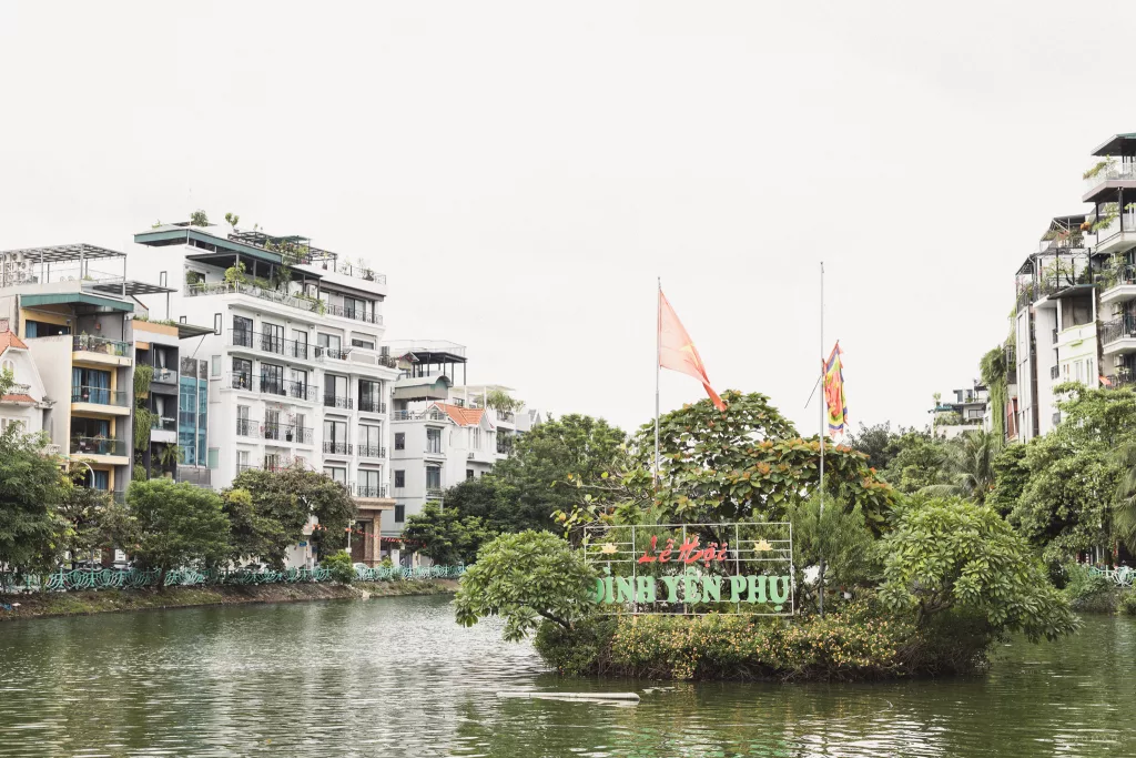 Schild auf einer bepflanzten Insel im Wasser mit der Beschriftung Dinh Yen Phu mit Gebäuden ringsherum