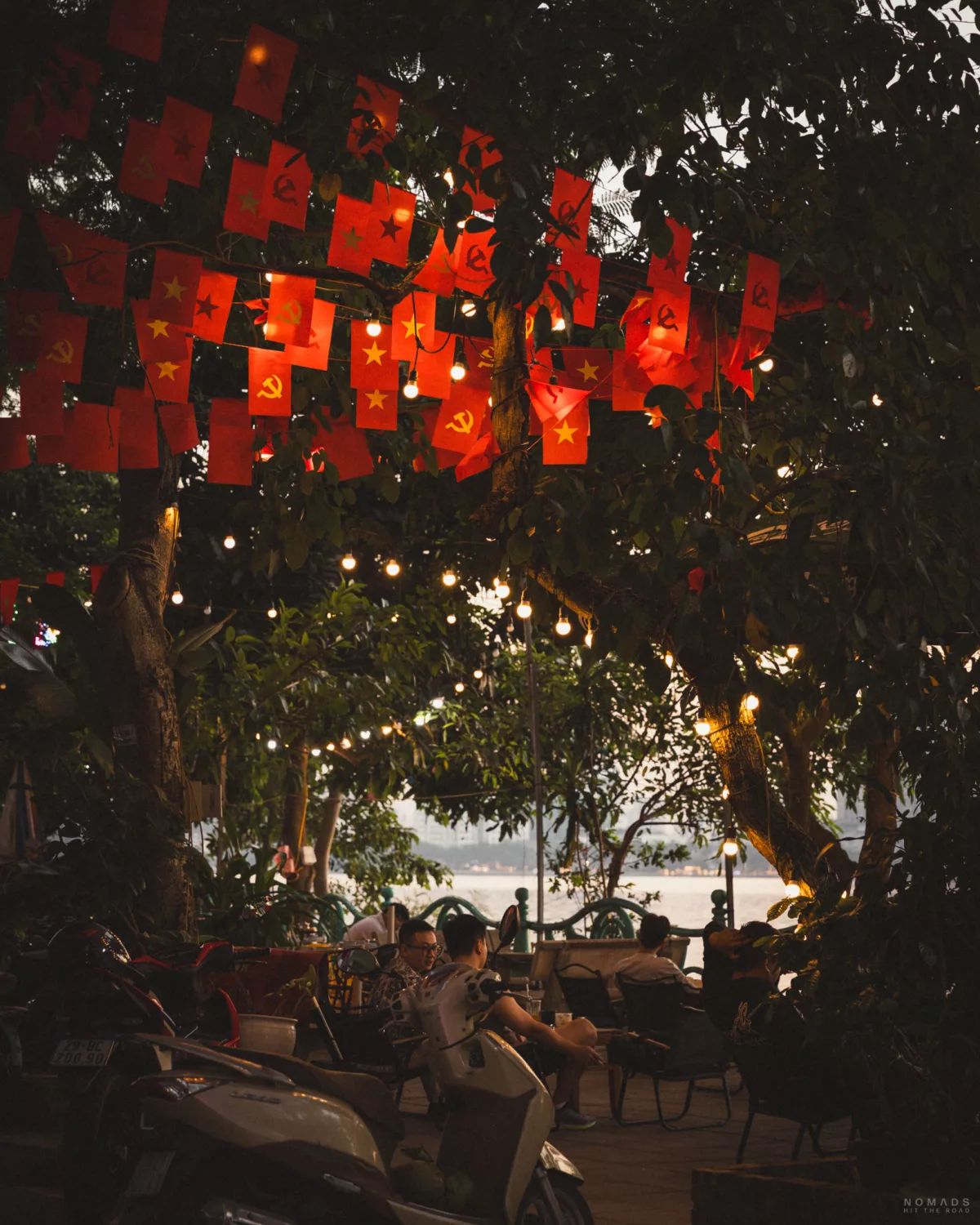 Menschen sitzen am frühen Abend bei Sonnenuntergang am West Lake in Tay Ho, während vietnamesische Fahnen in den Bäumen hängen