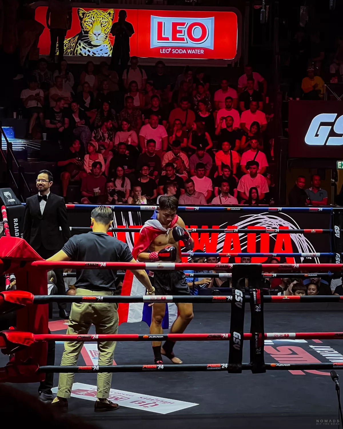 Muay Thai Kämpfer und weitere Menschen im Ring im Rajadamnern Stadion, im Hintergrund Menschen auf den Plätzen und Werbung von Leo