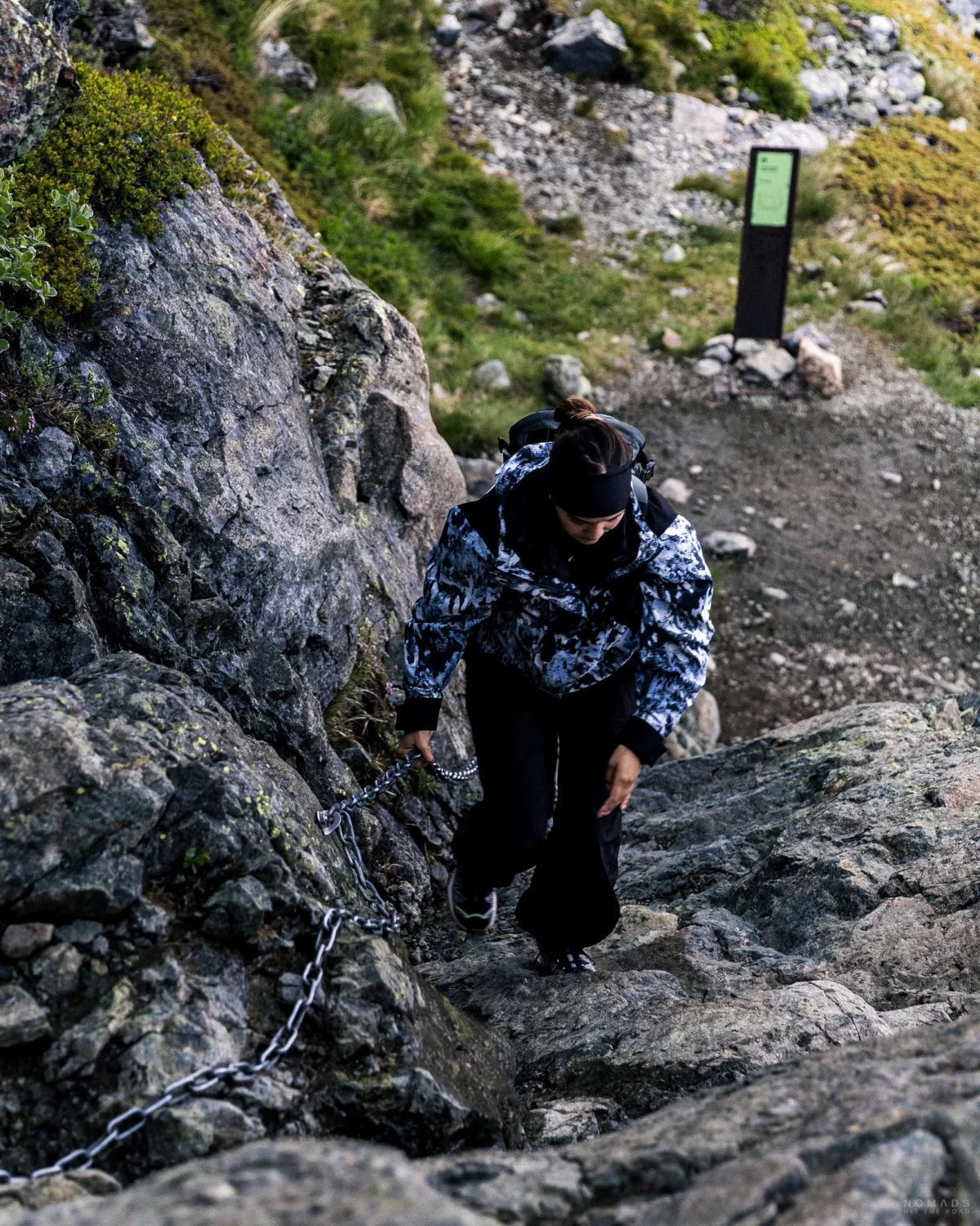Frau in Regenjacke und mit Rucksack auf dem Rücken steigt eine Felswand hoch, an der Bergkante hängt eine Metallkette zum festhalten