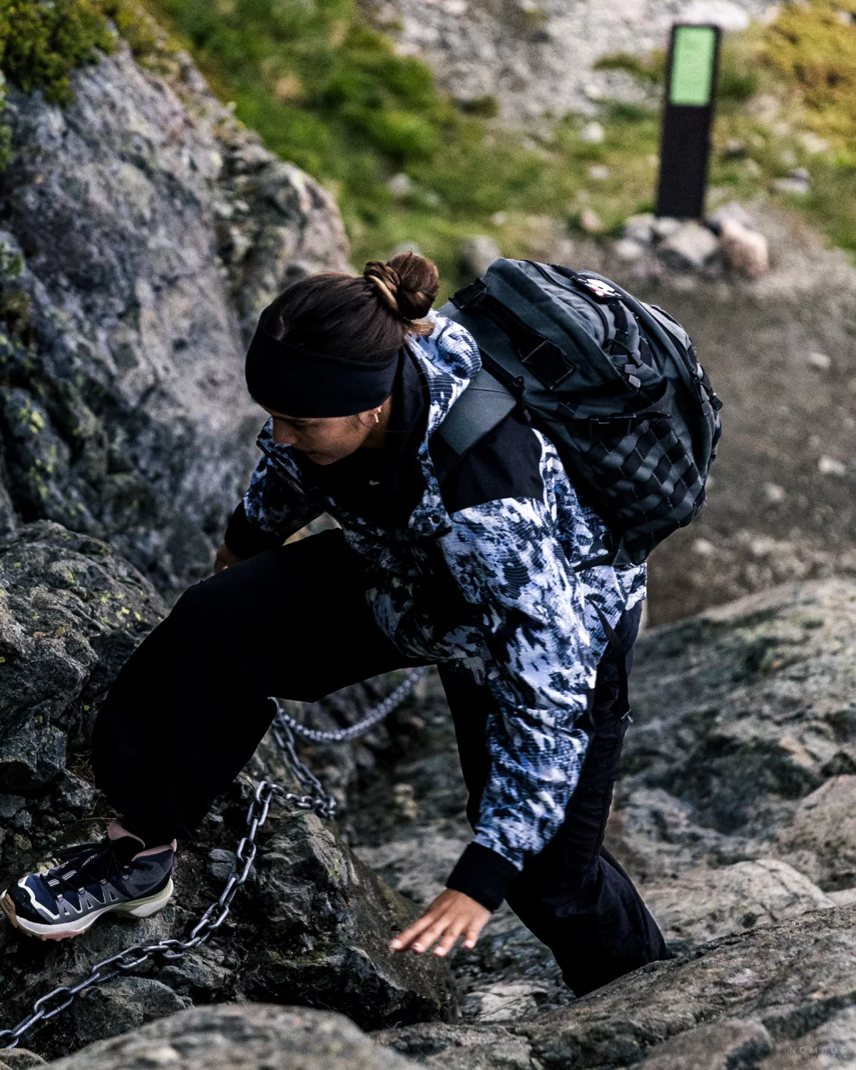 Frau in Regenjacke und mit Rucksack auf dem Rücken steigt eine Felswand hoch, an der Bergkante hängt eine Metallkette zum festhalten