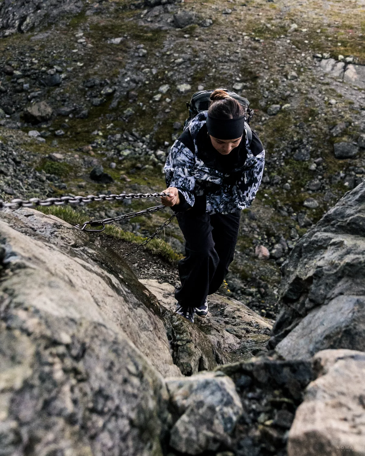 Frau in Regenjacke und mit Rucksack auf dem Rücken steigt eine Felswand hoch, an der Bergkante hängt eine Metallkette zum festhalten