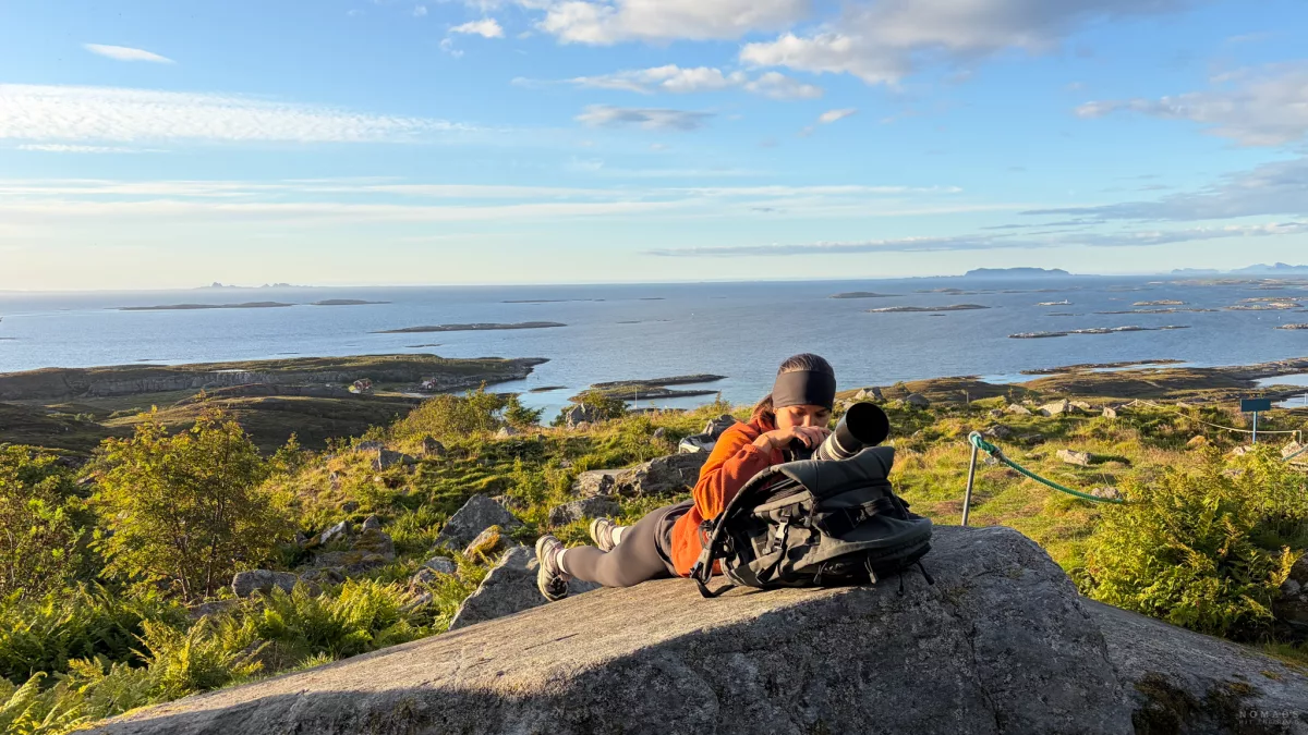 Frau liegt auf einem Felsen und fotografiert, Kamera liegt auf einem Rucksack, Blick auf den Fjord und Inseln im Hintergrund
