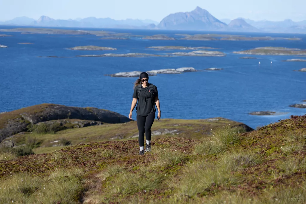 Frau wandert auf einem grünen Berg, im Hintergrund Fjord und Berge