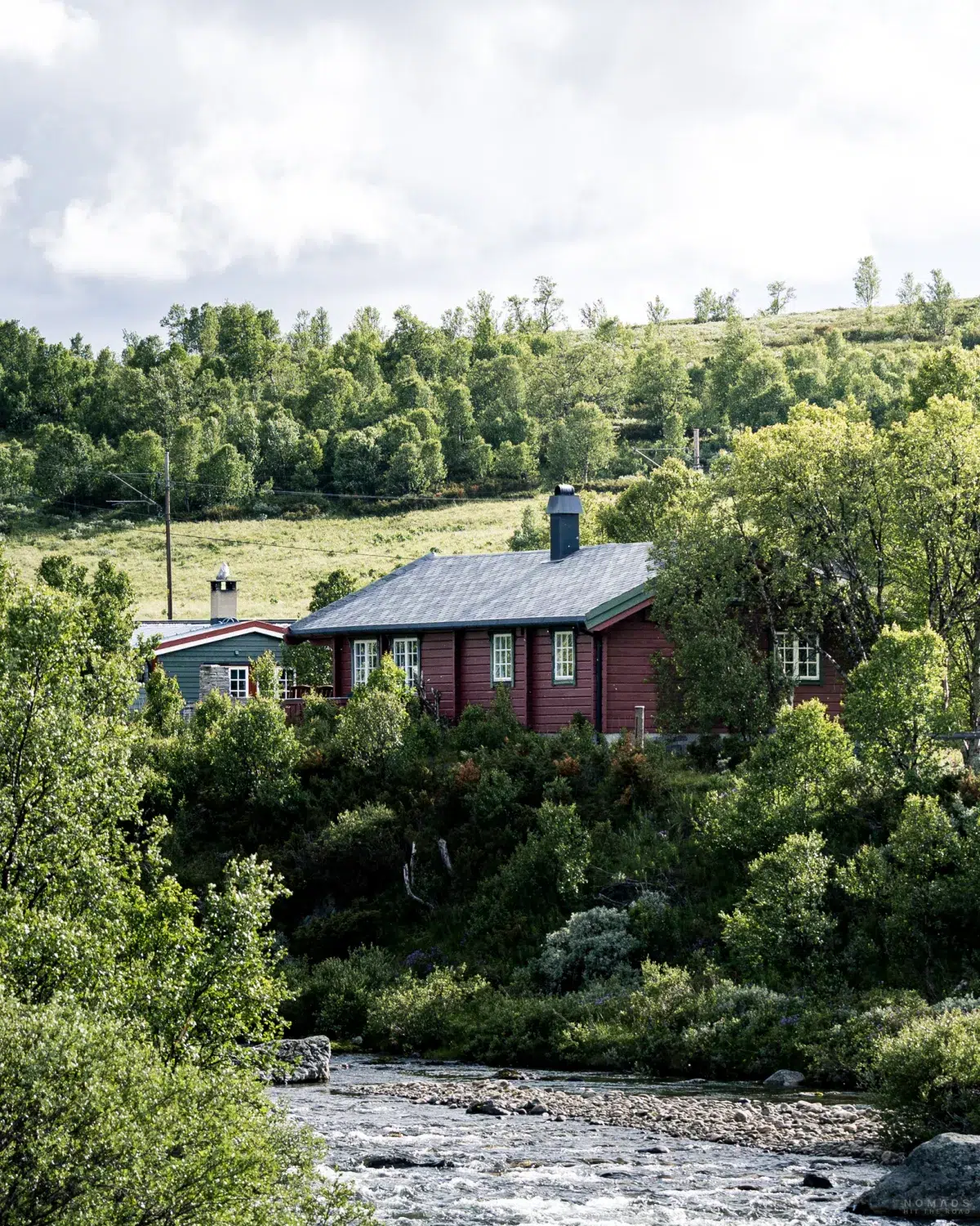 Blick auf ein Holzhaus zwischen Bäumen und Büschen an einem Fluss gelegen im Dovrefjell