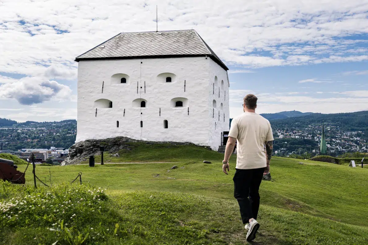 Mann läuft auf einer grünen Wiese auf die weiße Festung Kristiansten in Trondheim zu, dahinter liegt die Stadt