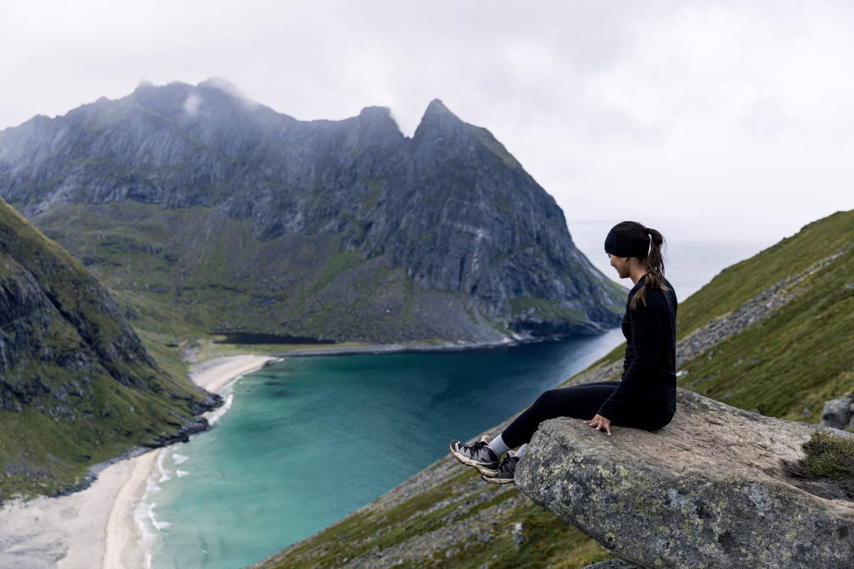 Frau sitzt auf einem Stein am Aussichtspunkt mit Blick auf den Kvalvika Beach auf den Lofoten sowie einer Bergekette im Hintergrund