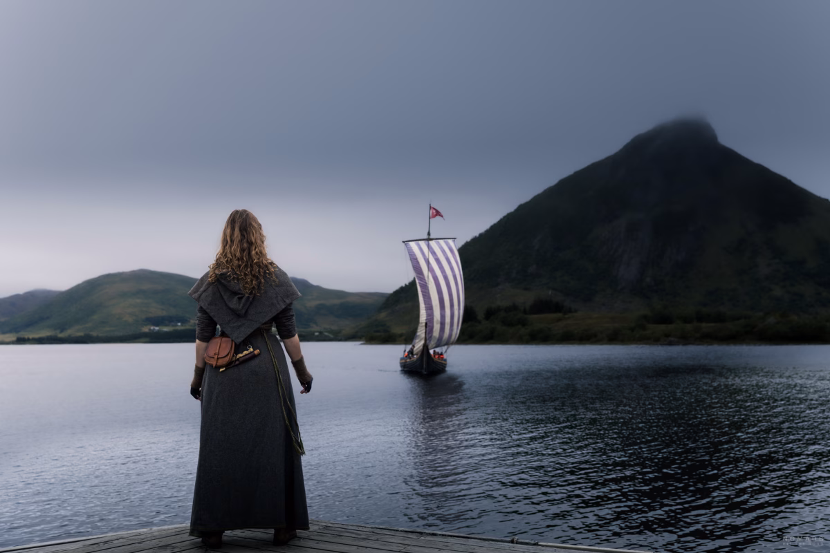 Person im Wikingermuseum und Kostüm steht am Ufer und wartet auf ein Segelschiff, im Hintergrund ein Berg im Nebel