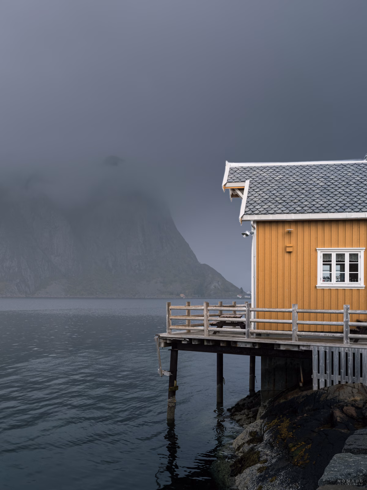 Gelbes Haus steht am Fjord, im Hintergrund Berge im Nebel